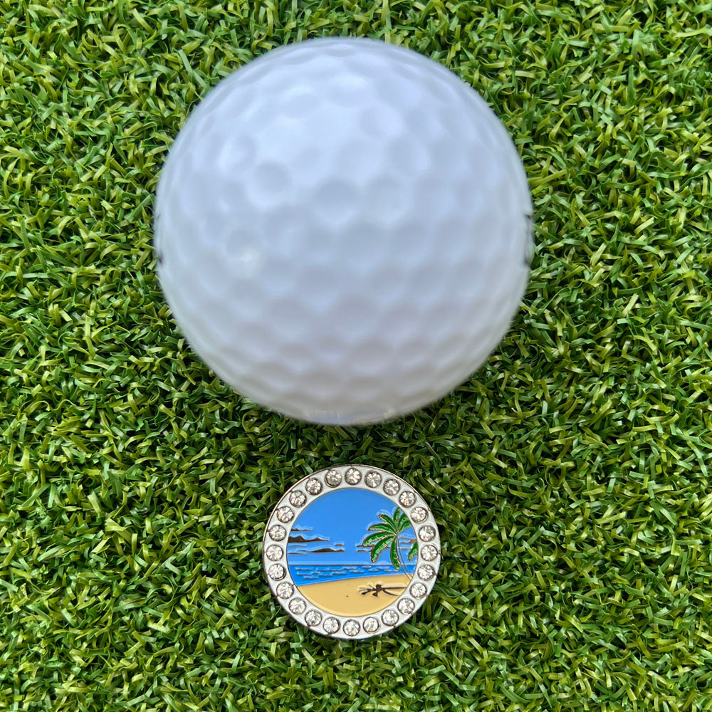 Golf ball on green grass with a golf ball marker featuring a beach design.