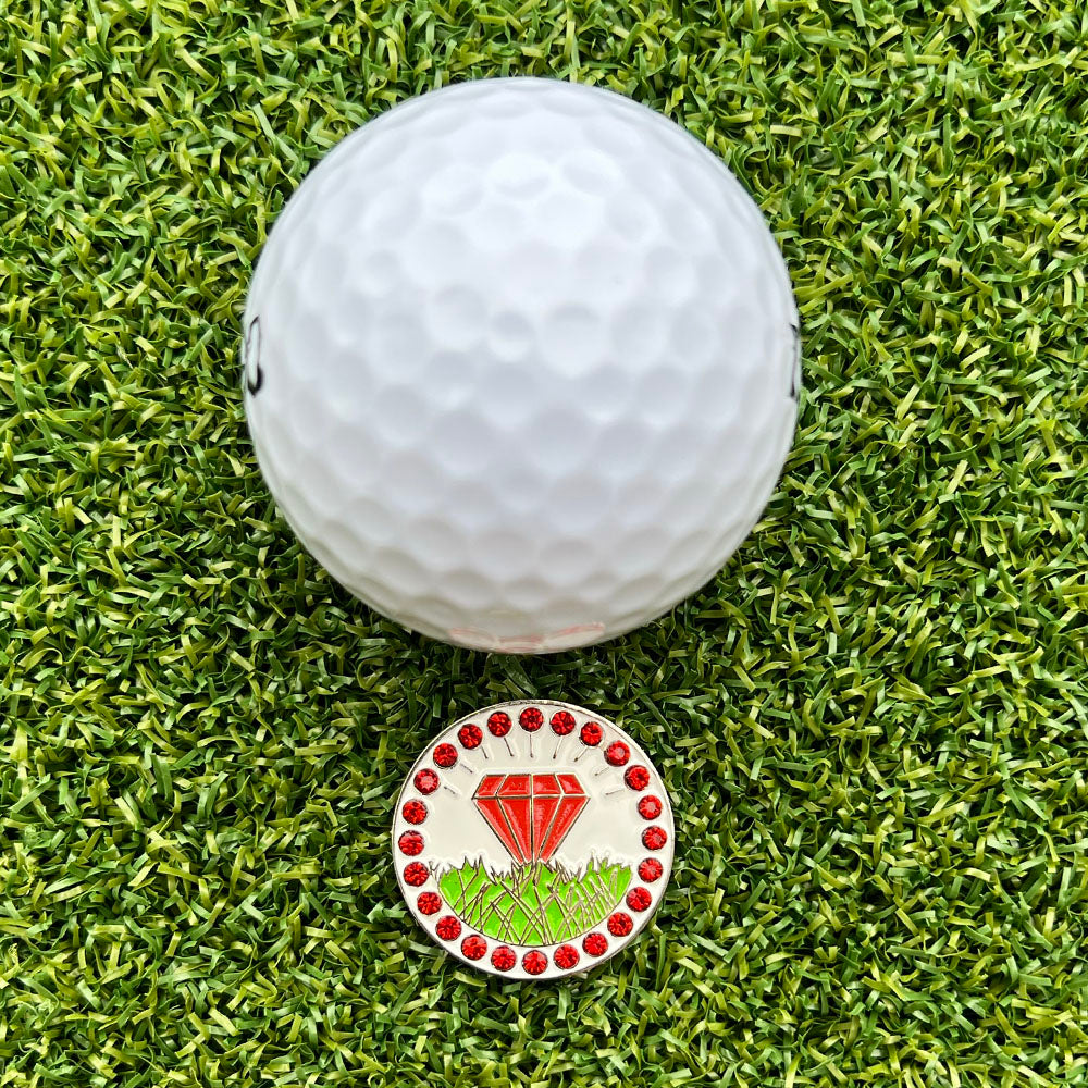 Golf ball on grass with a golf ball marker featuring a red diamond design.