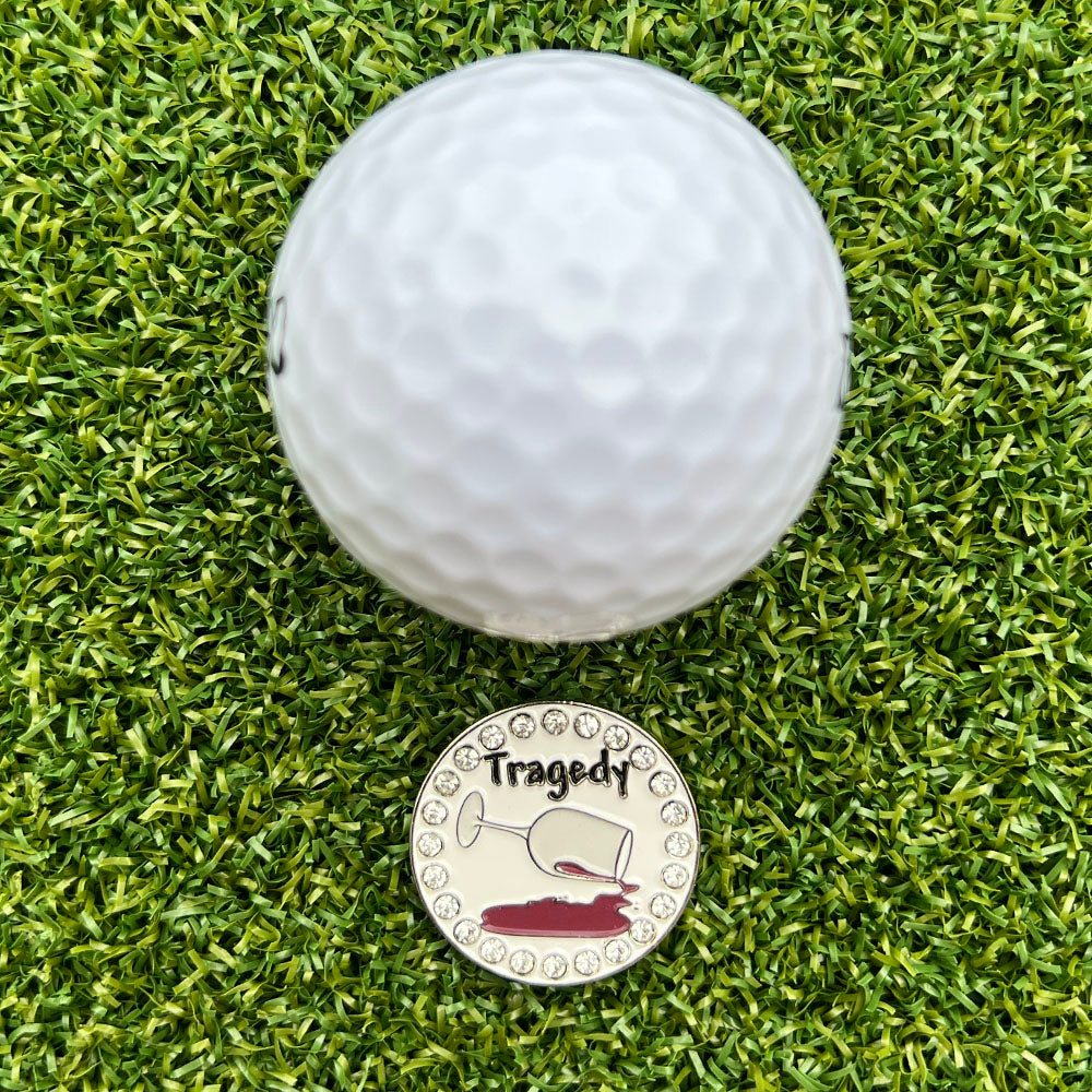 Golf ball on grass with a metal golf ball marker with wine and says 'Tragedy'.