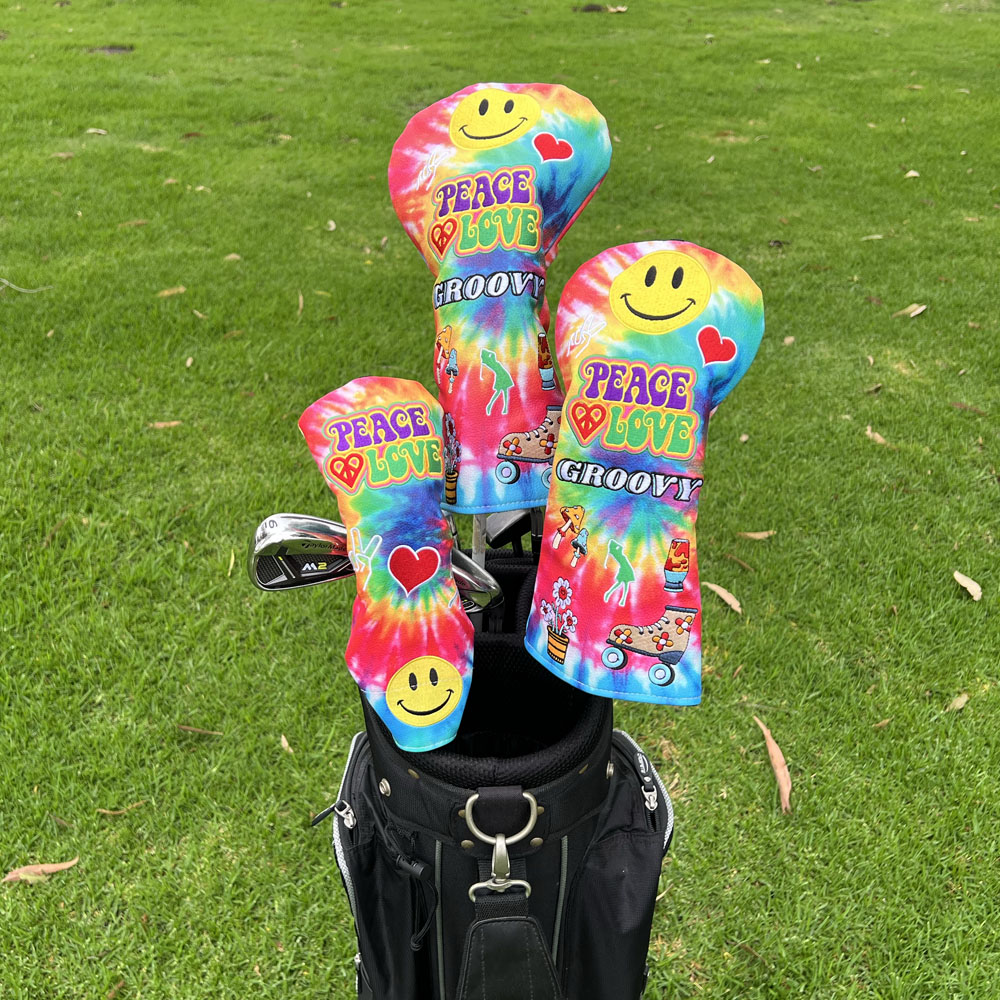 Three colorful tie-dye golf club headcovers with smiley faces and the words 'PEACE LOVE GROOVY' are shown in a golf bag on a grassy fairway