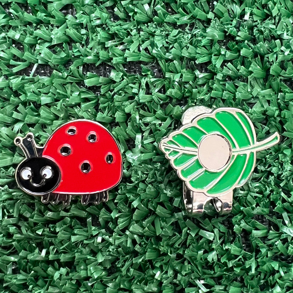 red ladybug golf ball marker next to a leaf shaped magnetic hat clip, on grass background