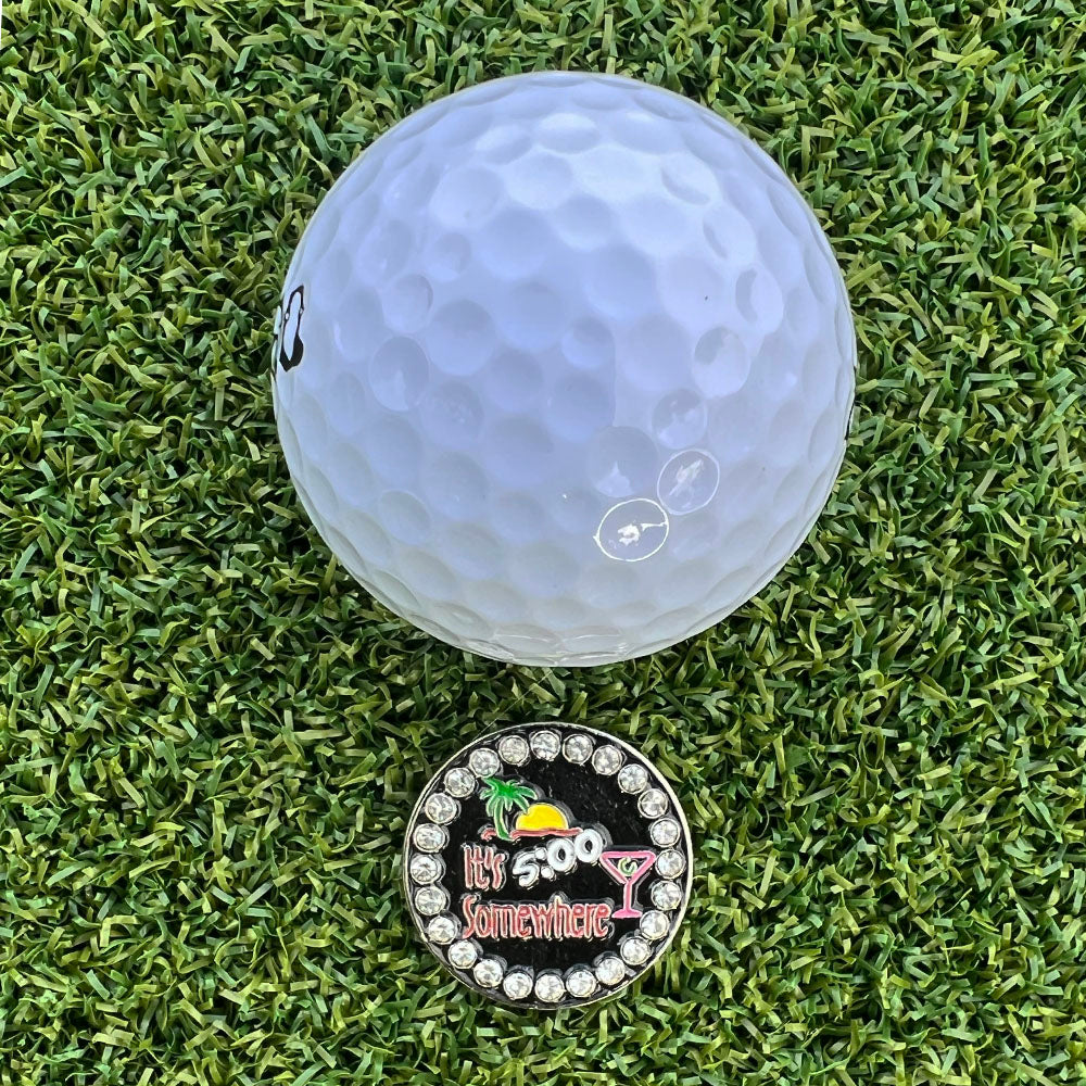 Golf ball on a grassy surface with a colorful 5 O'clock Somewhere golf ball marker below.