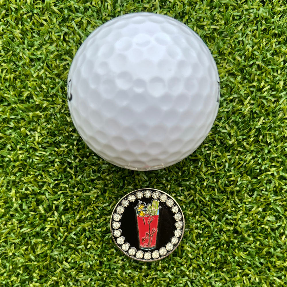 Golf ball on a green grass surface with a golf ball marker featuring a bloody mary drink