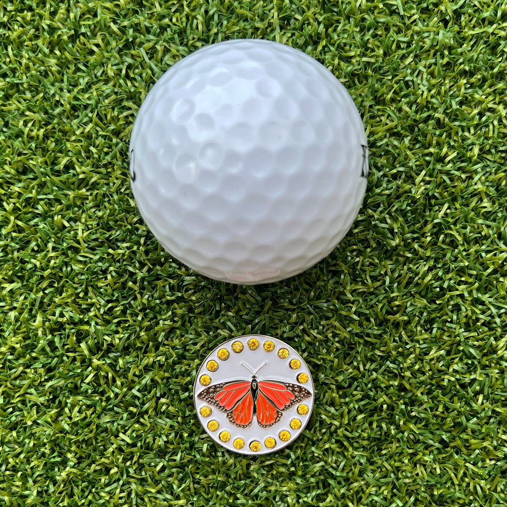 Golf ball on green grass with a butterfly-themed golf ball marker.