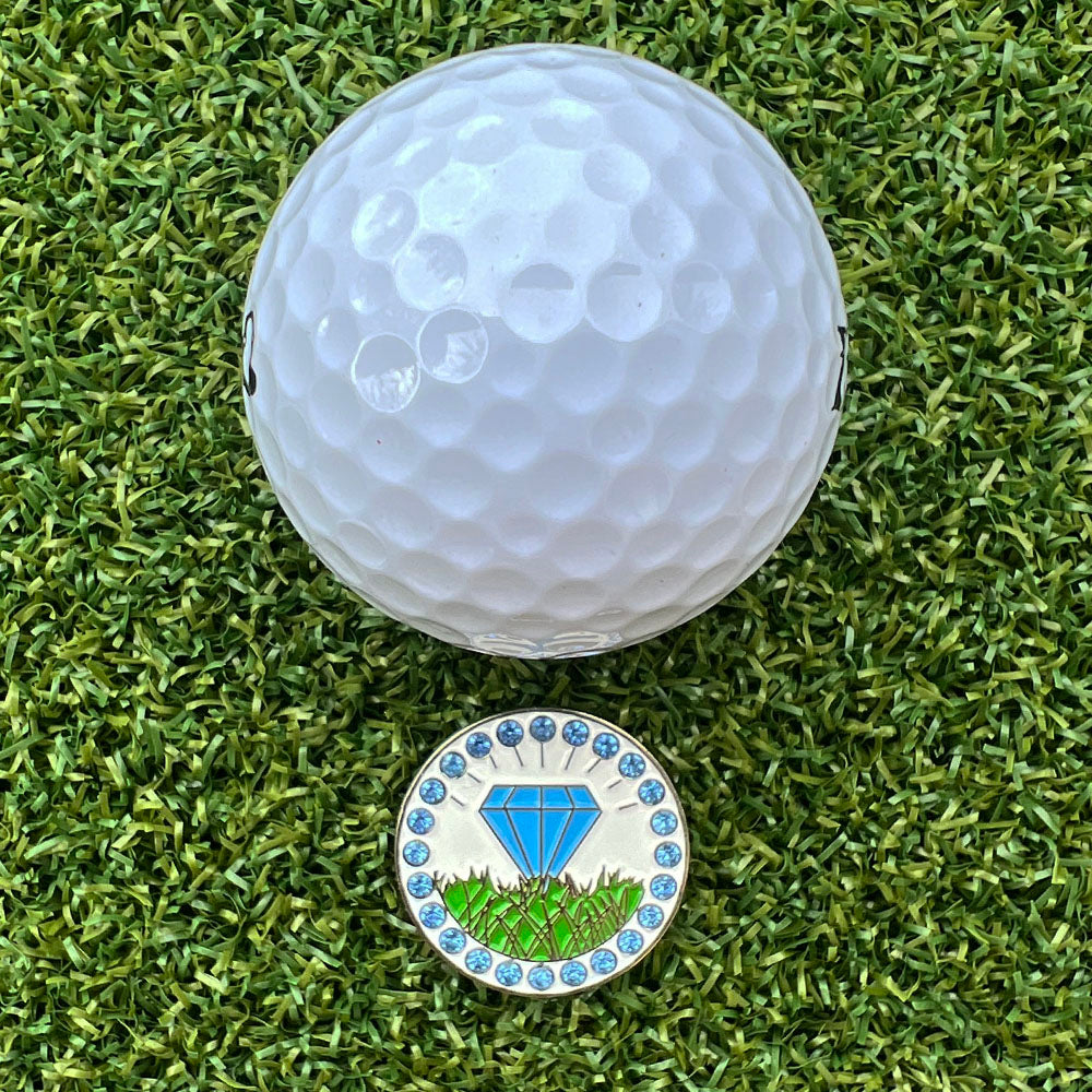 Golf ball on grass with a golf ball marker featuring a blue diamond design.