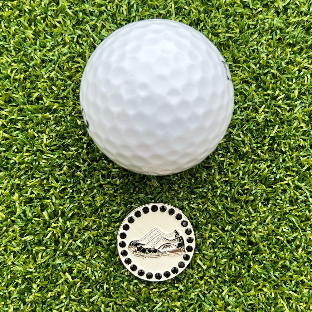 Golf ball on a green grass surface with a ball marker featuring a pair of black and white golf shoes.