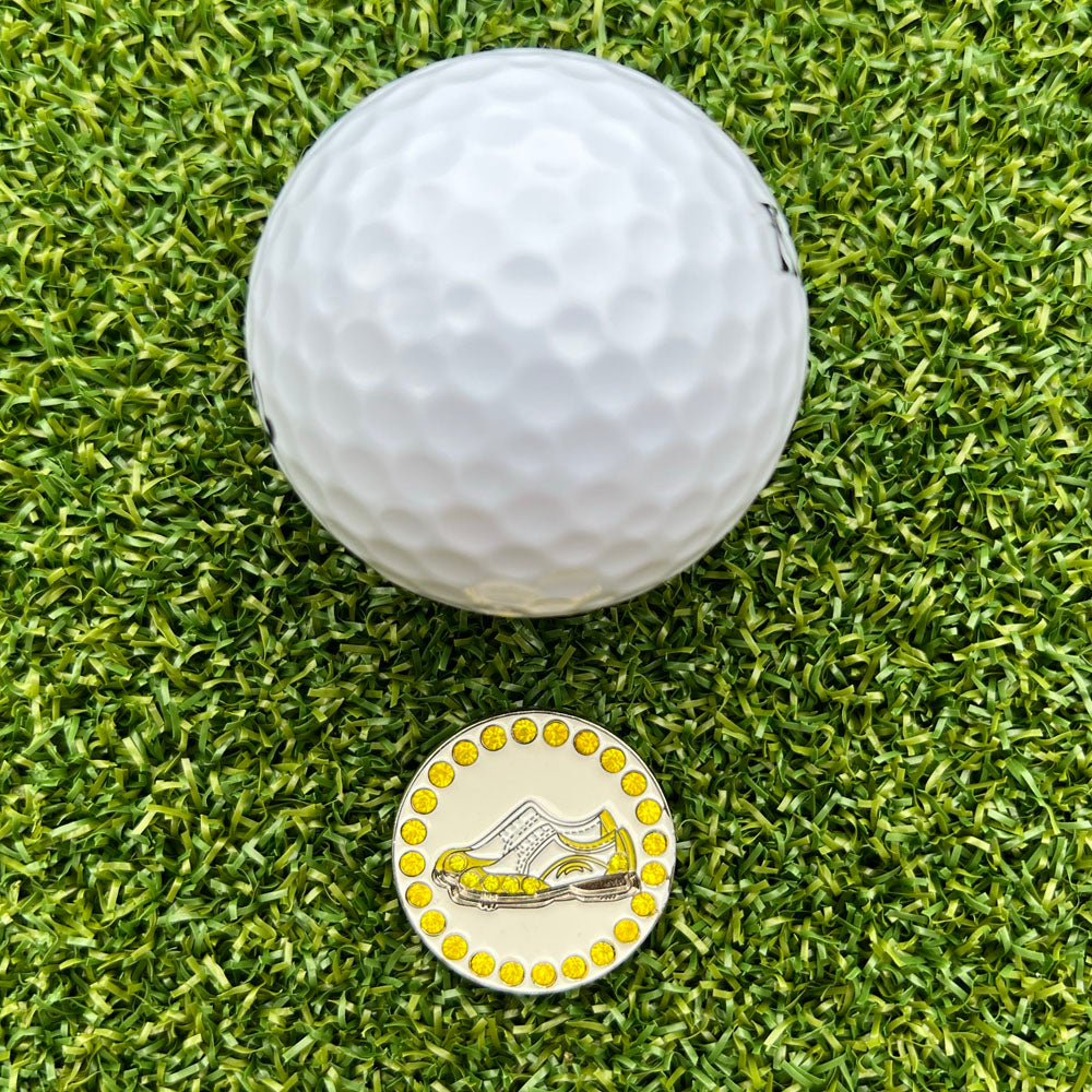 Golf ball on grass with a golf ball marker featuring a yellow and white shoe design.