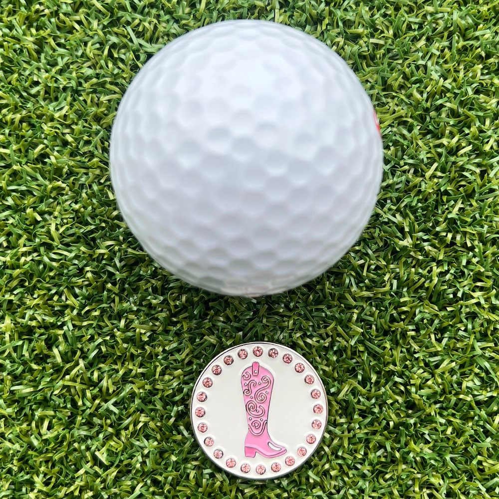 White golf ball on green grass with a pink western boot design golf ball marker.