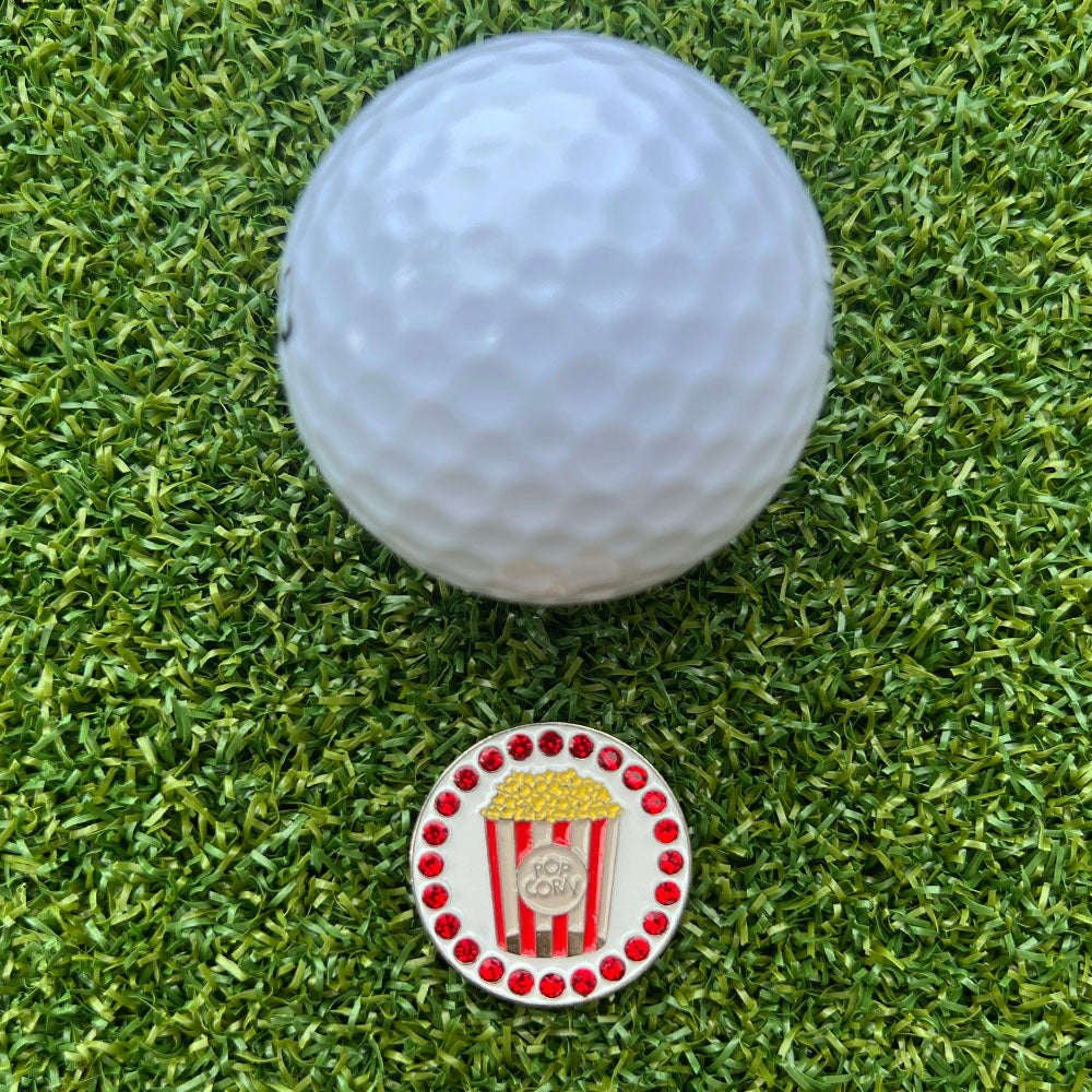 Golf ball on grass with a decorative golf ball marker featuring popcorn bucket design.