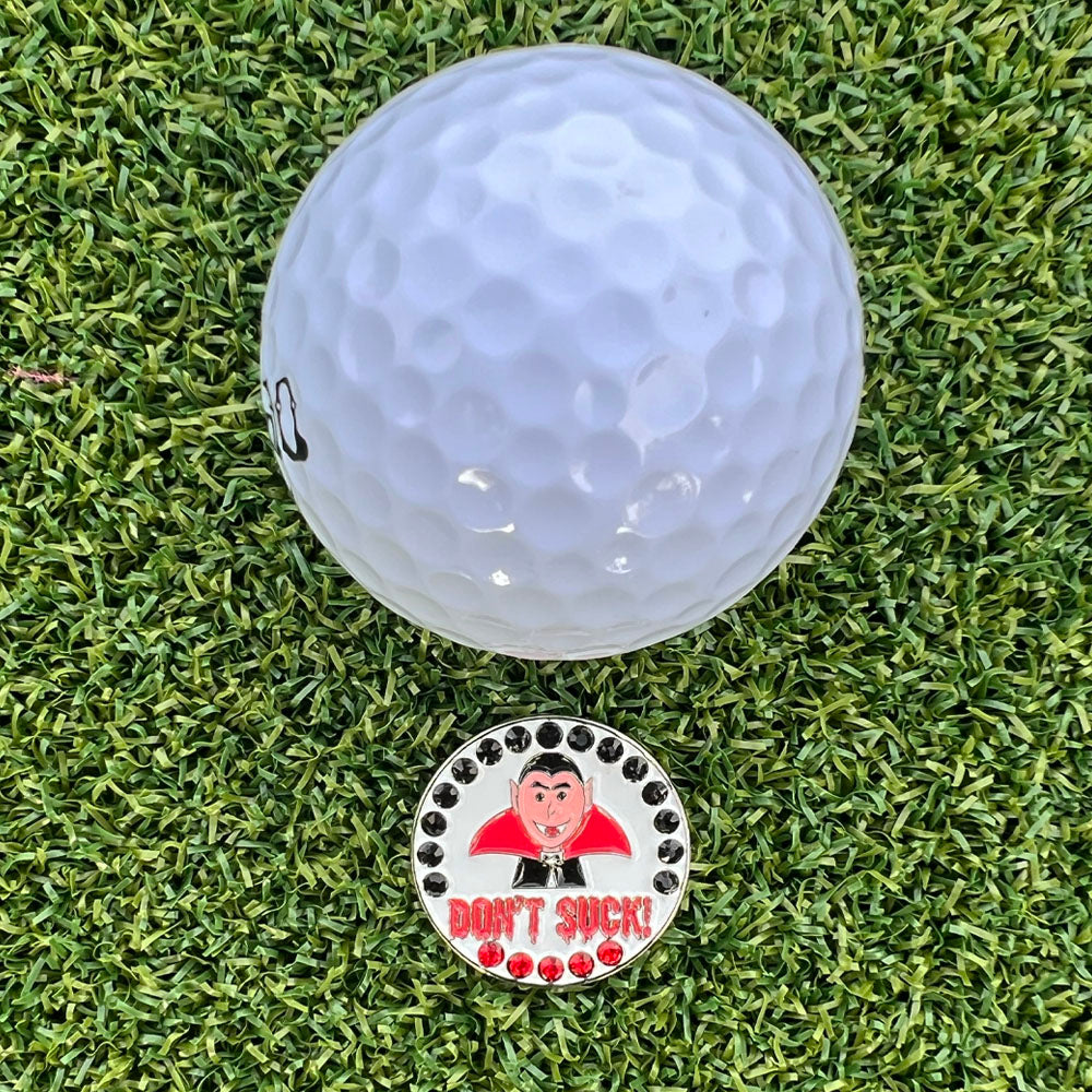 Golf ball on a green grass surface with a humorous golf ball marker featuring a vampire and 'Don't Suck!' text.