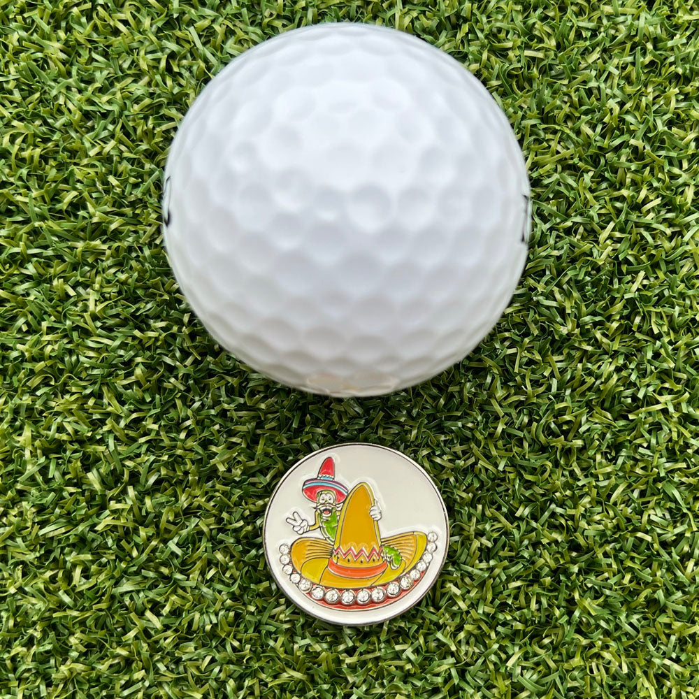 Golf ball with a colorful golf ball marker featuring a sombrero & worm design on grass.