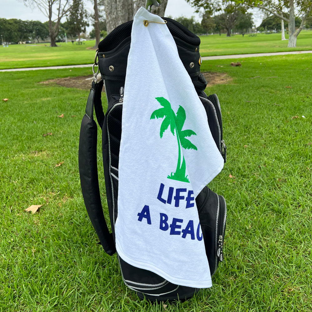 Golf bag with a towel featuring palm trees and 'Life's A Beach' text on grass.