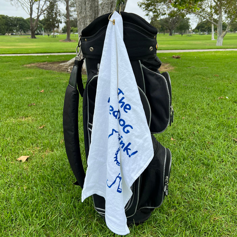 Golf bag with a white towel hanging out, featuring blue text and a dog with a drink, on a grassy background.