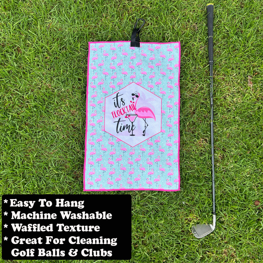 Golf towel with flamingo design and text on grass, next to a golf club.
