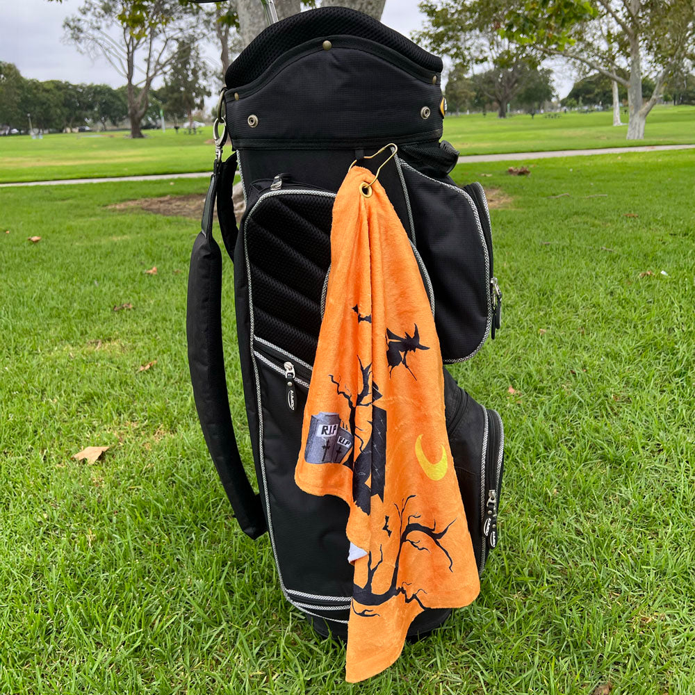 Black golf bag with an orange towel featuring Halloween designs on a grassy background