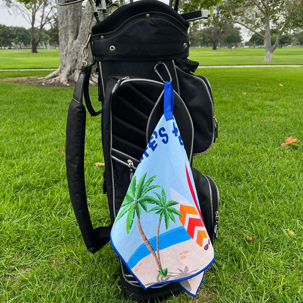Golf bag with a towel featuring palm trees and a beach scene on a grassy background